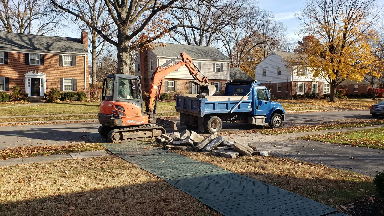 Mini excavator loading broken driveway concrete into a dump truck on a quiet Olathe residential street with ground-protection mats on the lawn