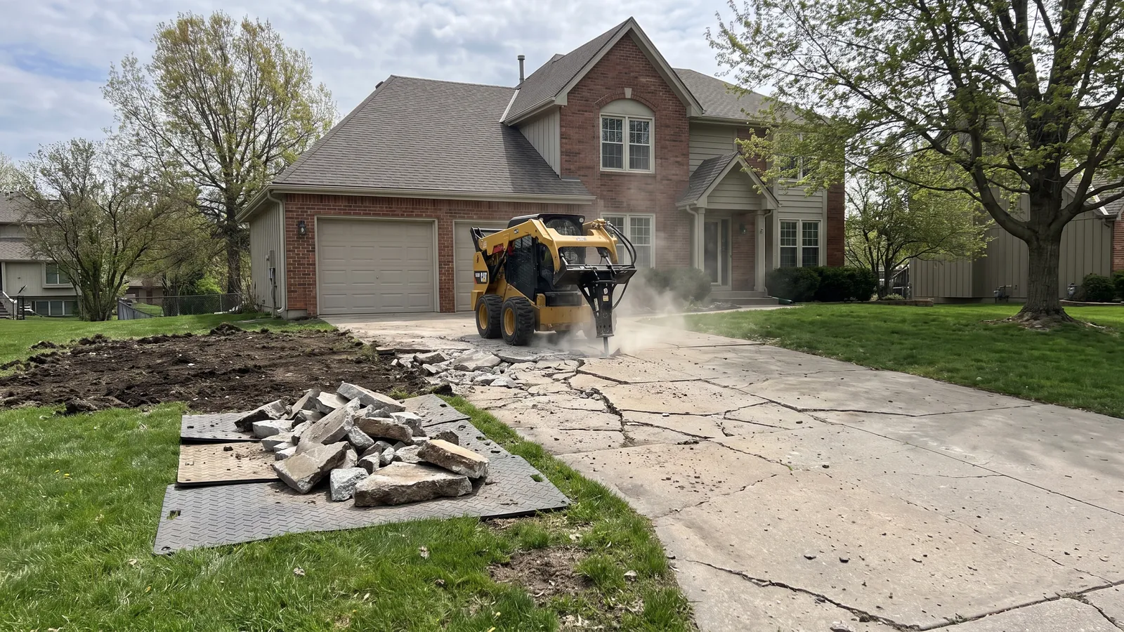 Skid steer breaking out a cracked and heaved 1990s concrete driveway at a two-story home in Olathe, Kansas, broken slabs piled for hauling
