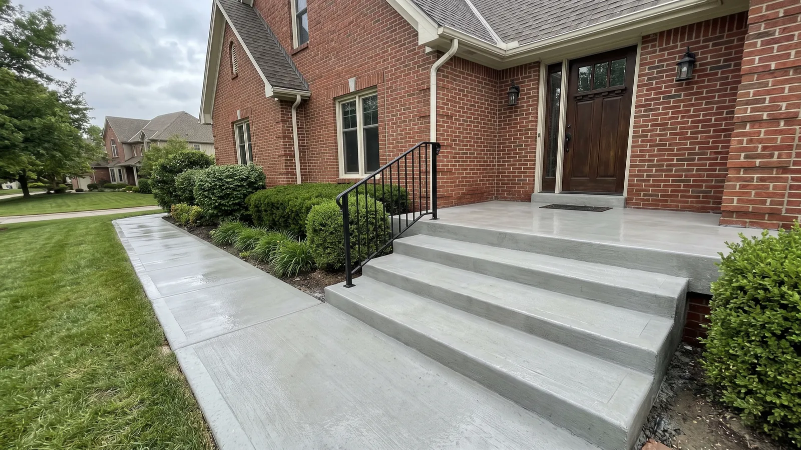 New concrete front stoop, three wide steps and a broom-finish walkway with a black metal handrail at a brick-front Olathe home