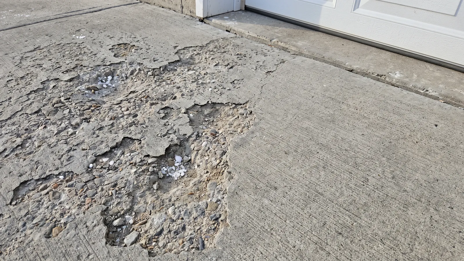 Close-up of a concrete driveway surface scaled and flaking from road salt and freeze-thaw, beside an intact broom-finished section